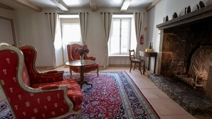 Una habitación con chimenea, sillones tapizados en rojo, una mesa redonda con un florero y una alfombra estampada.
