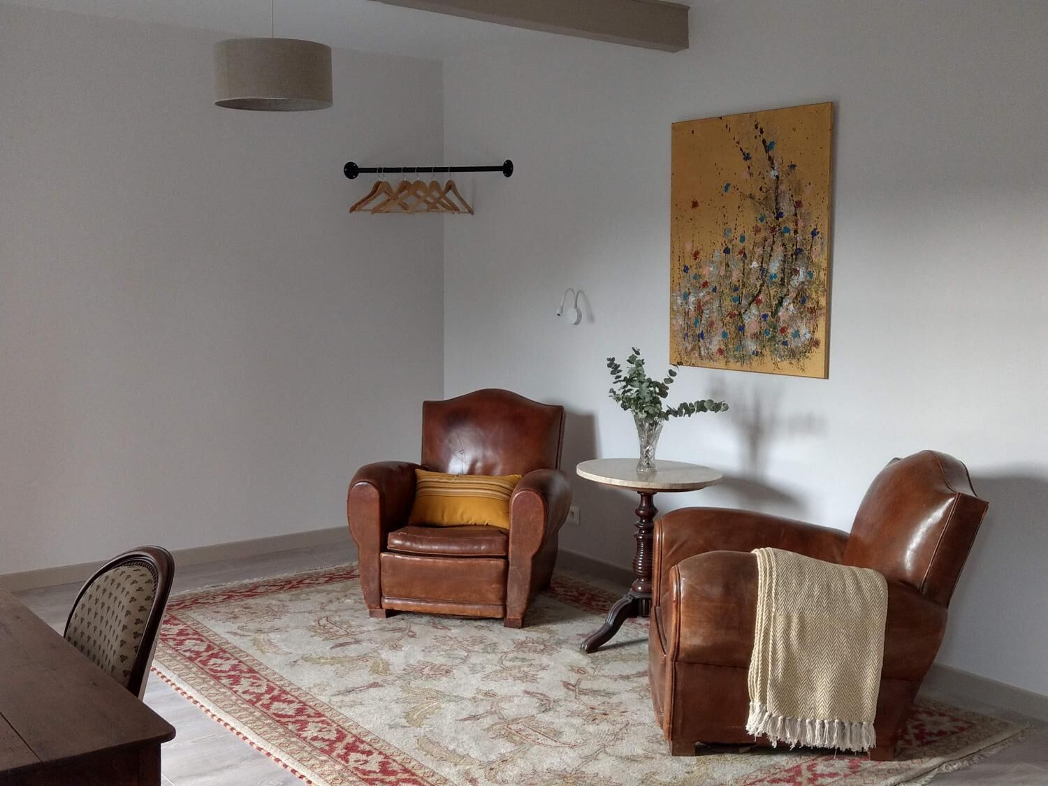 Un salón con un sillón de cuero marrón, una mesita redonda con un florero, una alfombra estampada y un cuadro en la pared.