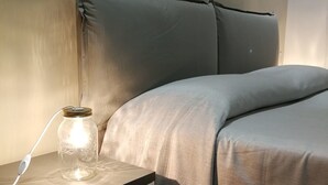 A neatly made bed with a grey headboard and pillows, a bedside table with a lamp and a jar, and a drawer unit. - B&B Napoli Culture (Naples)