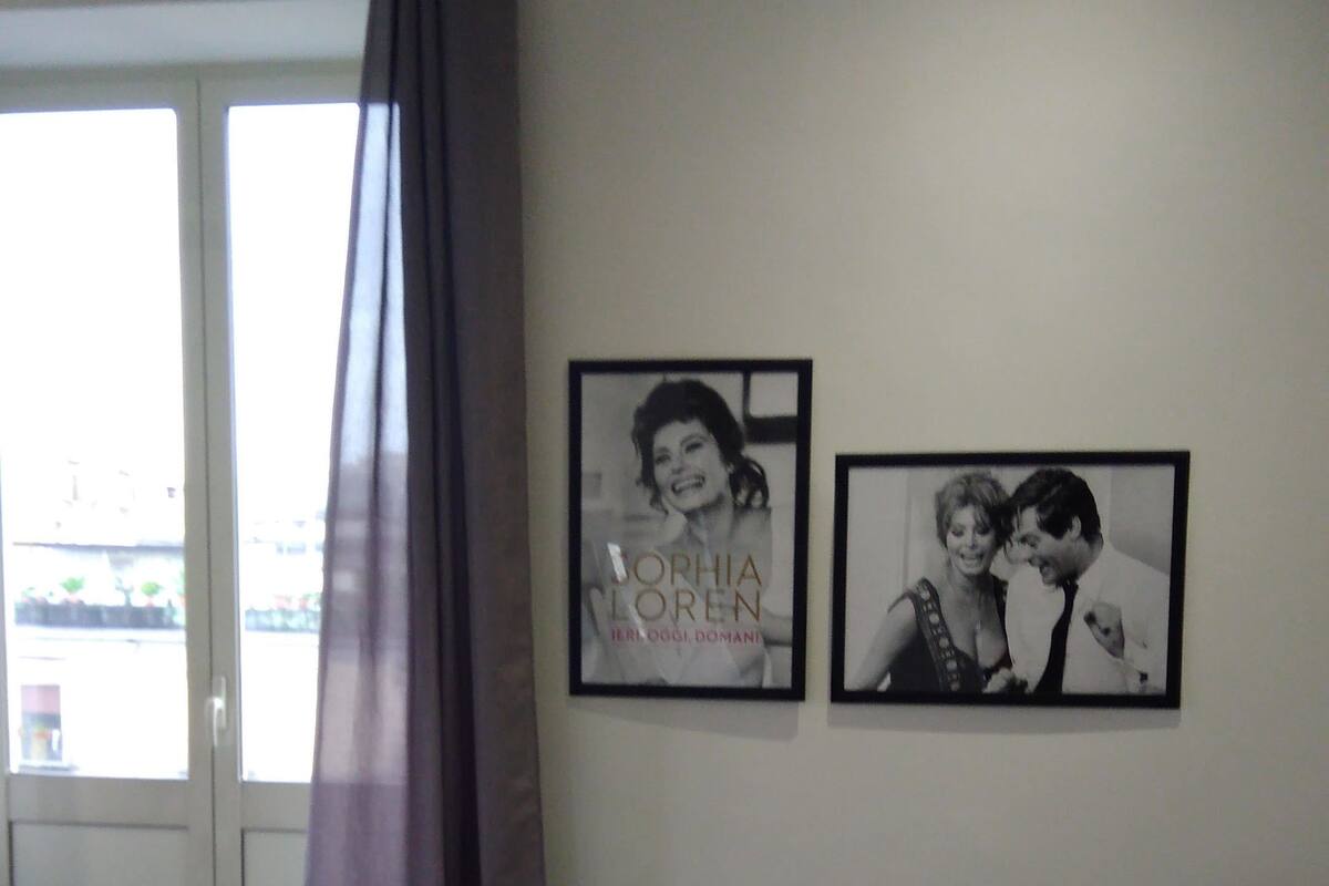 A hotel room with a bed, two framed pictures on the wall, a window with curtains, and a view of the outside.