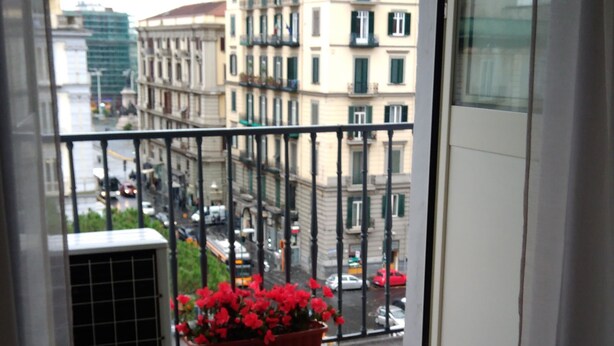 A balcony with a table set for breakfast, a potted plant with red flowers, and a view of buildings in the background. - B&B Napoli Culture (Naples)