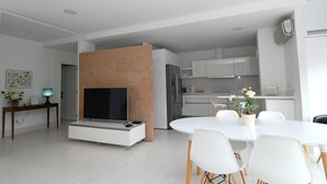 A modern living room with a flat-screen TV, a dining table with chairs, a kitchen area with cabinets, and a small table with a lamp and flowers.