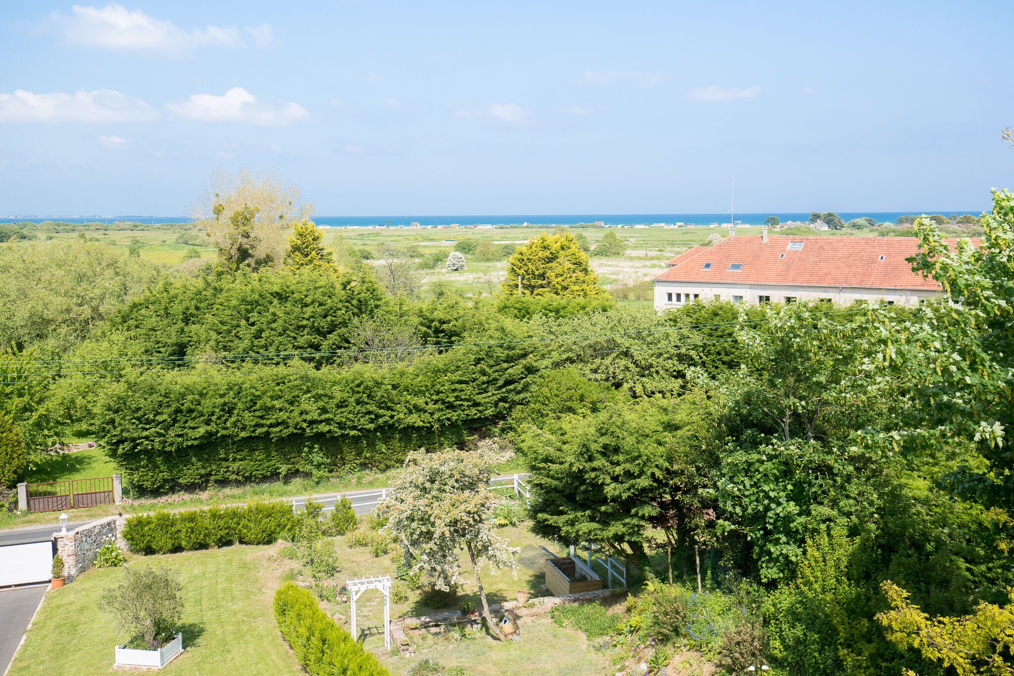 A residential area with a house featuring a red roof, surrounded by lush greenery and a clear view of the sea.