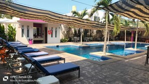 Un área de piscina con sillones de descanso y sombrillas, un pequeño edificio con una puerta rosa y palmeras.