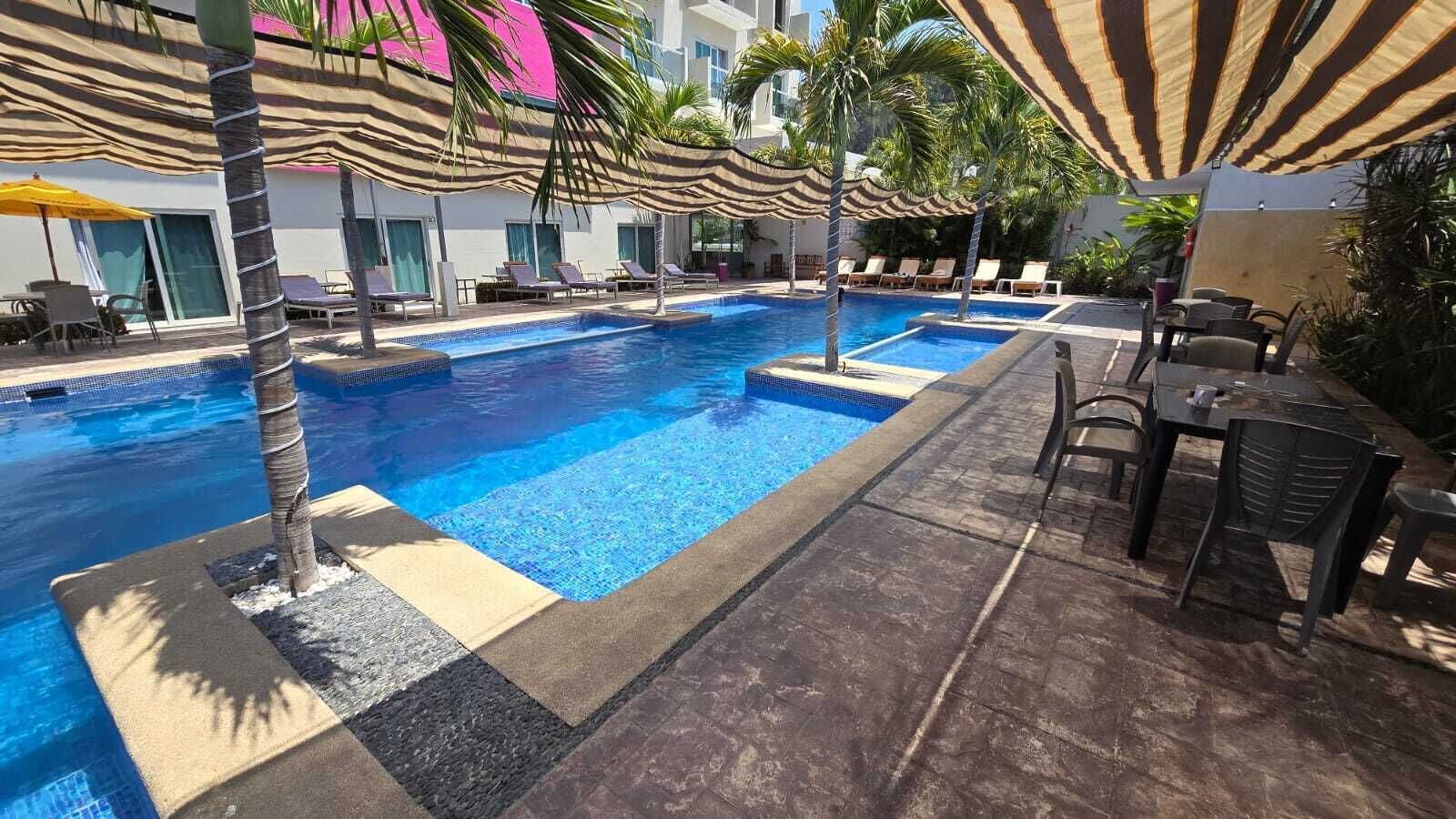 Ein Pool mit Liegestühlen und Sonnenschirmen, umgeben von Palmen und einem Gebäude mit großen Fenstern.
