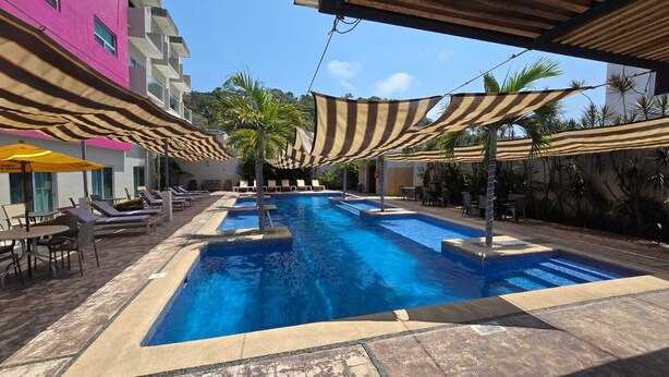 Piscina com espreguiçadeiras e guarda-sóis, cercada por edifícios e palmeiras.