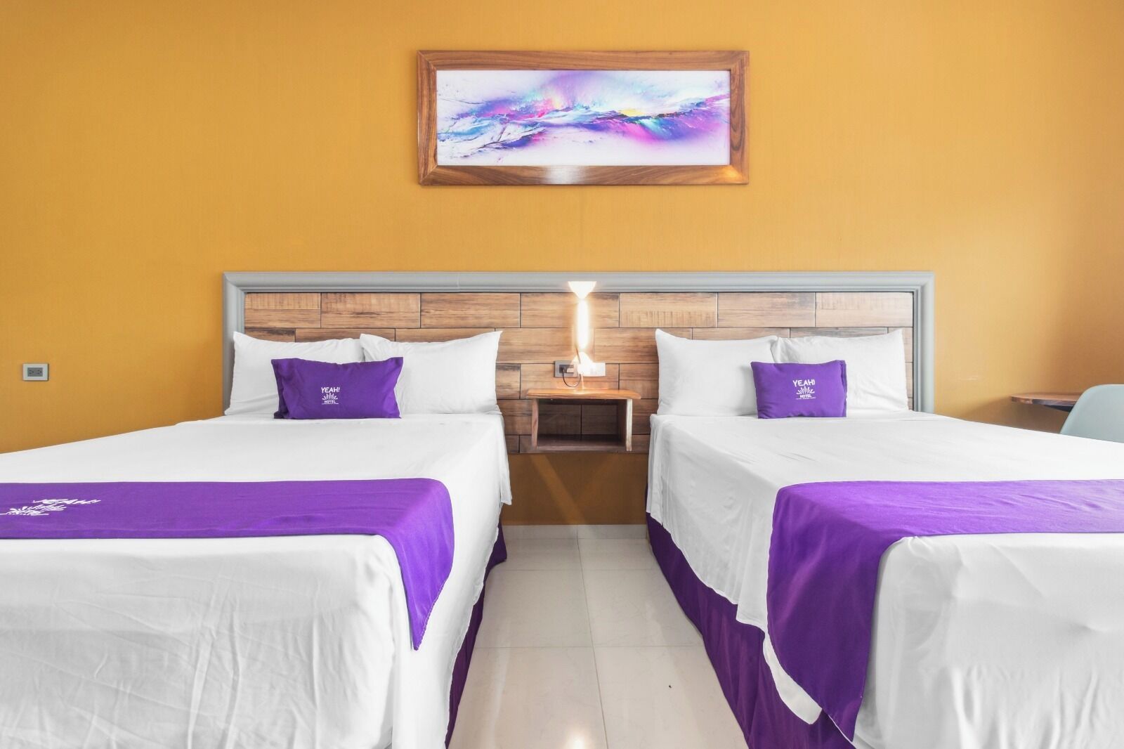 Habitación de hotel con dos camas, cada una con sábanas blancas y detalles morados, un cabecero de madera, una mesita de noche con una lámpara y un cuadro abstracto colorido en la pared.