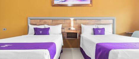 Habitación de hotel con dos camas, cada una con sábanas blancas y detalles morados, un cabecero de madera, una mesita de noche con una lámpara y un cuadro abstracto colorido en la pared.