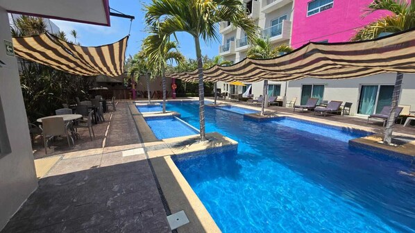 Una piscina con sillones de playa y sombrillas, rodeada de palmeras y un edificio con fachada rosa.