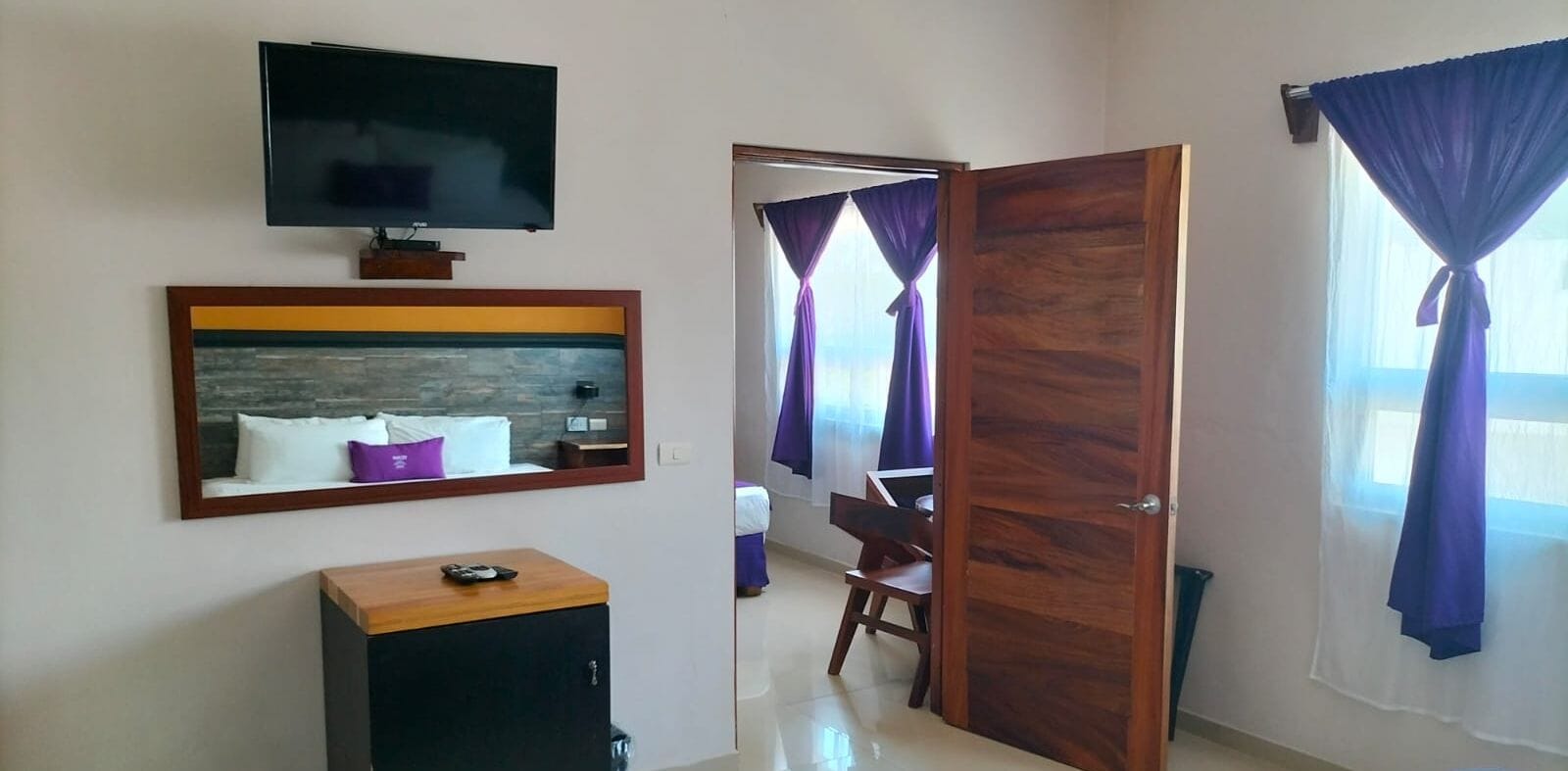 A hotel room with a television, a framed artwork, a wooden cabinet, a door, and a window with curtains.