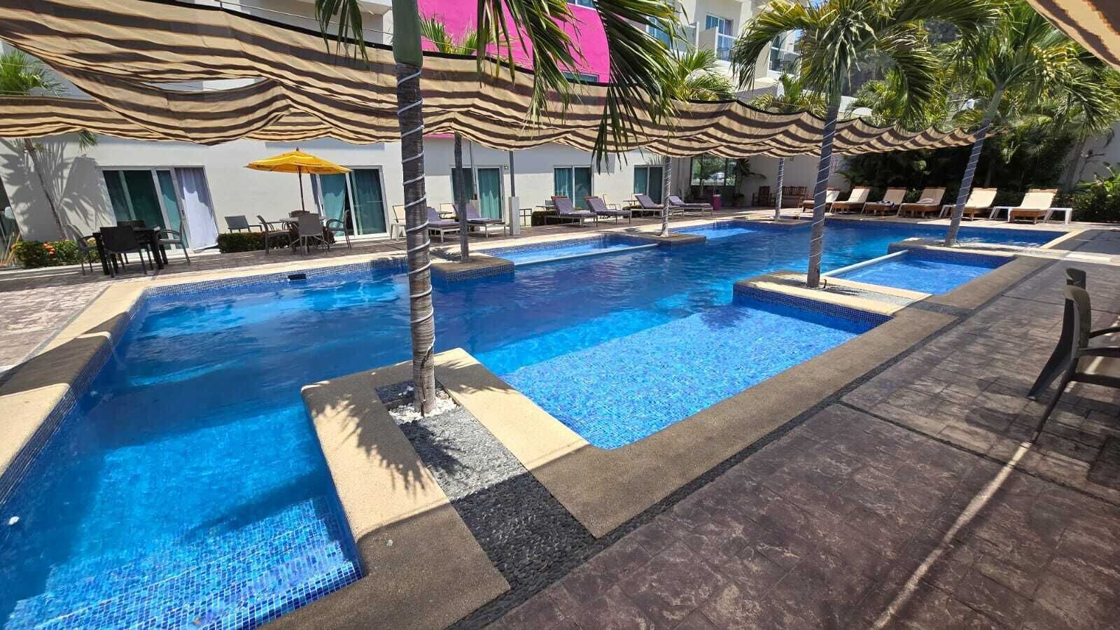 Une piscine entourée de palmiers et d’un bâtiment doté de grandes fenêtres, avec des chaises longues et des parasols à proximité.