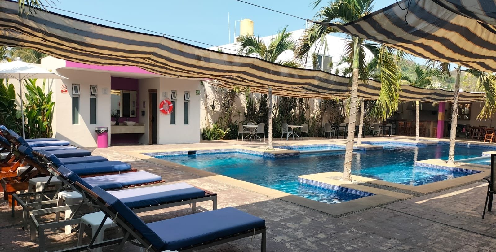 A pool area with lounge chairs and umbrellas, a small building with a pink door, and palm trees.