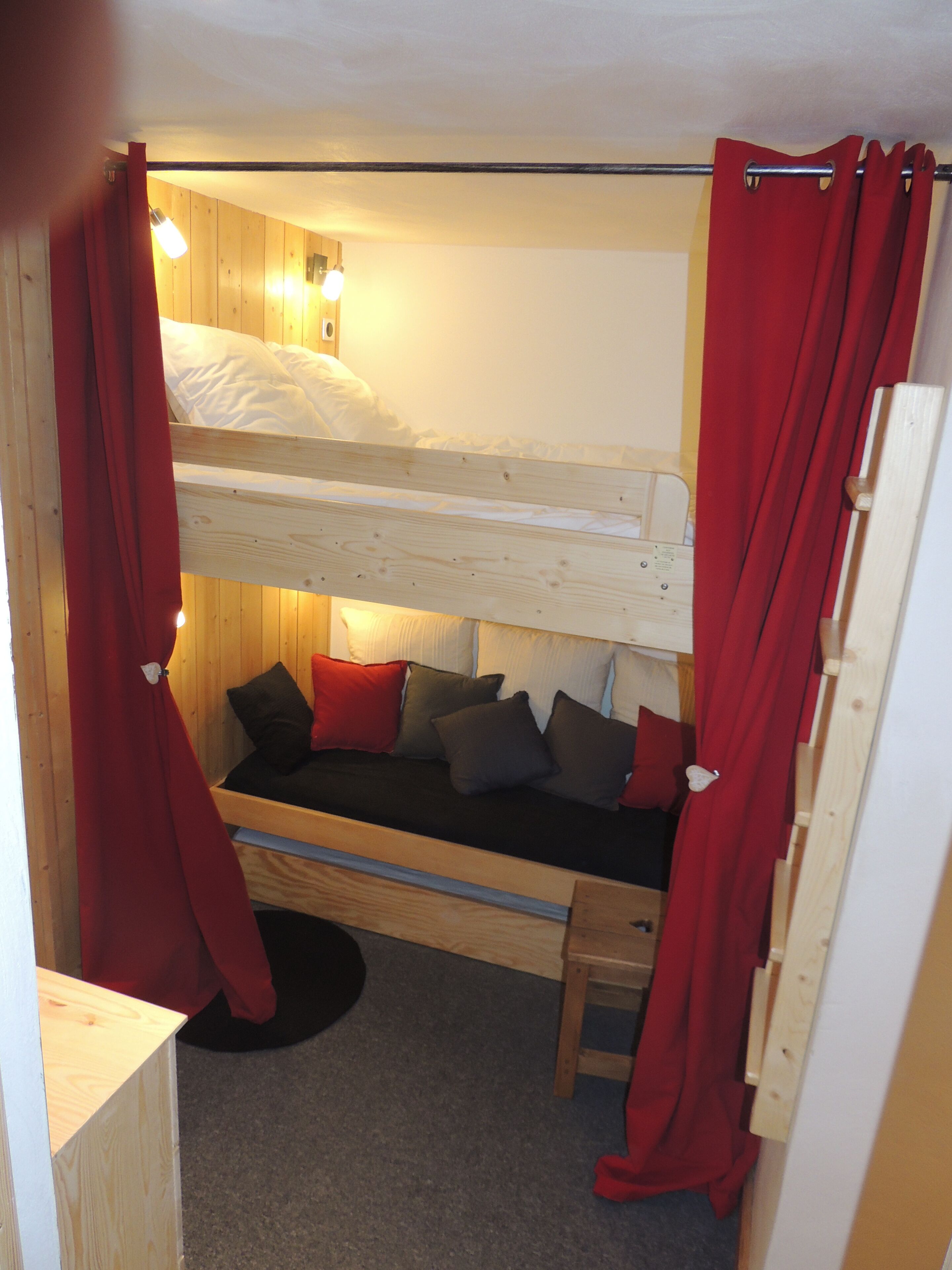 Dormitorio con literas, suelo de madera, un taburete pequeño y cortinas rojas.