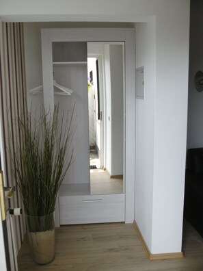 Um corredor com uma planta em vaso, uma porta branca e um guarda-roupa aberto com roupas penduradas.