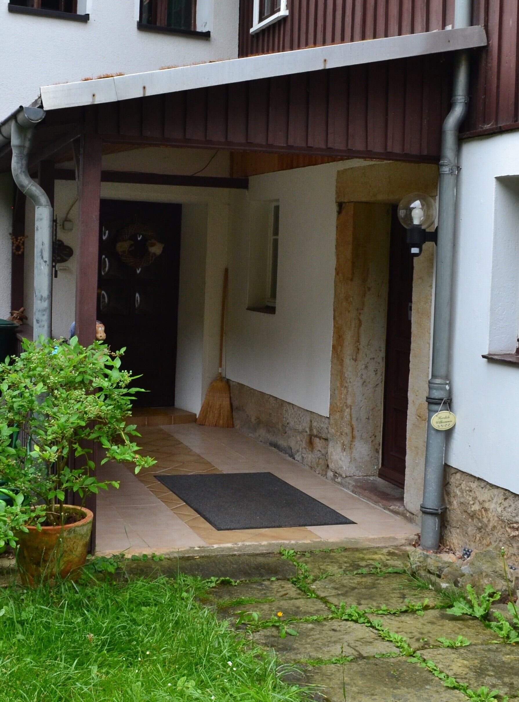 A covered entrance to a building with a wooden door, a broom leaning against the wall, and a potted plant in the foreground.
