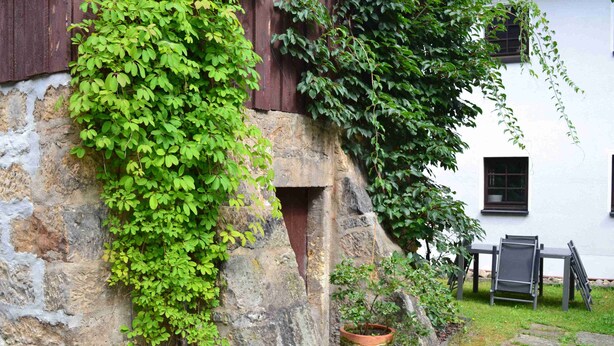 Un muro de piedra cubierto de hiedra, una planta en maceta y una mesa con sillas en un jardín.