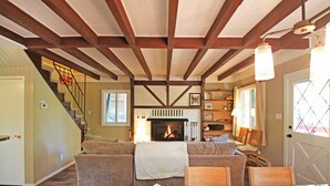 A cozy living room with a fireplace, wooden beams, a staircase, and a bookshelf.