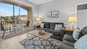 Un salón espacioso con un sofá gris, dos mesitas de noche con lámparas, una mesa de centro y una alfombra estampada. Un gran ventanal corredero da a un balcón exterior con sillas y vistas a una zona residencial.