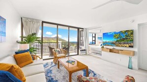 A modern living room with a sofa, armchairs, a coffee table, and a TV. There is a balcony with a view of the mountains.