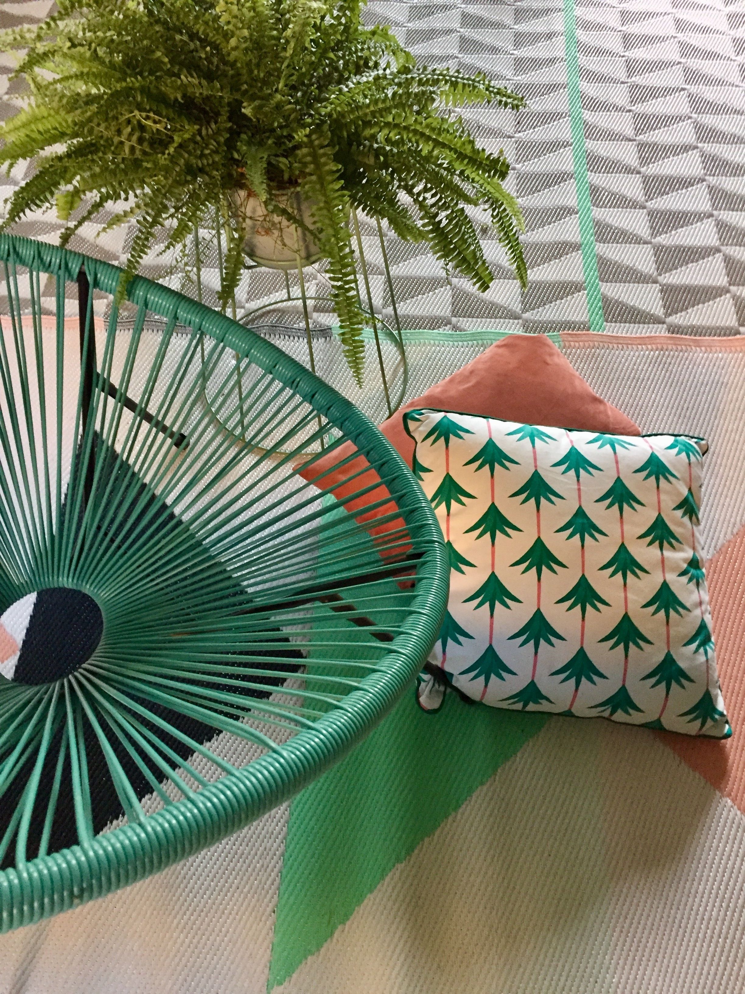 A green woven chair, a patterned cushion, and a plant in a room with a geometric patterned floor.