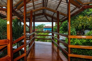A wooden bridge with railings leading to a covered area with a view of a blue building and lush greenery.