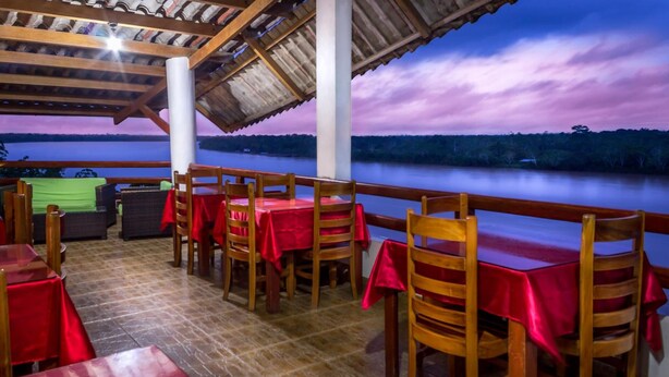 An outdoor dining area with wooden tables and chairs, a covered roof, and a view of a lake at dusk. - Hotel Rio Huallaga (Yurimaguas)