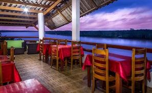 An outdoor dining area with wooden tables and chairs, a covered roof, and a view of a lake at dusk. - Hotel Rio Huallaga (Yurimaguas)