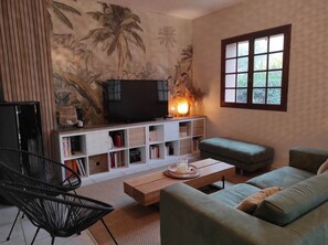 Una sala de estar con un televisor sobre un soporte de madera, una estantería, un sofá verde, una mesa de centro y una ventana con vistas a la vegetación.
