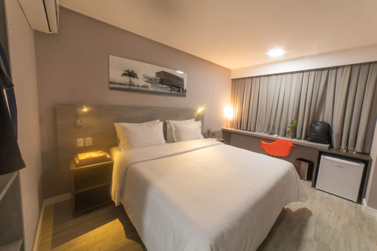 A hotel room with a bed, bedside table, nightstand, desk, chair, and a small refrigerator.