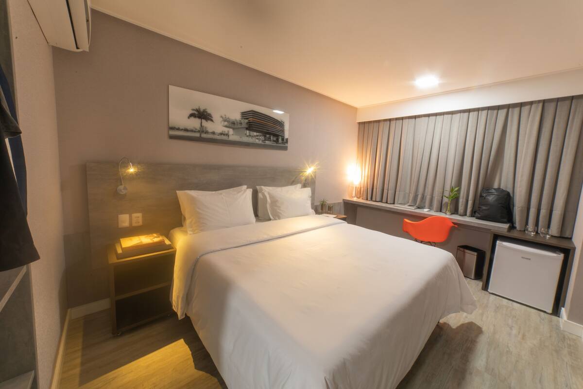 A hotel room with a bed, bedside table, nightstand, desk, chair, and a small refrigerator.
