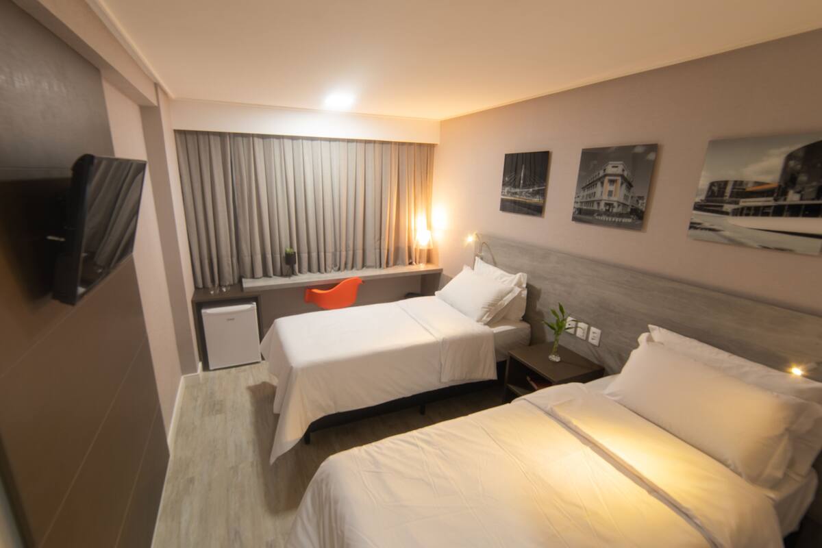 A hotel room with two beds, a desk, a chair, a television, and artwork on the walls.
