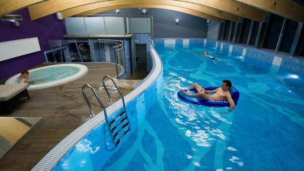 An indoor swimming pool with a wooden ceiling, a child in a small pool, and a person on a blue float. - Hotel Warmia Spa (Braniewo)