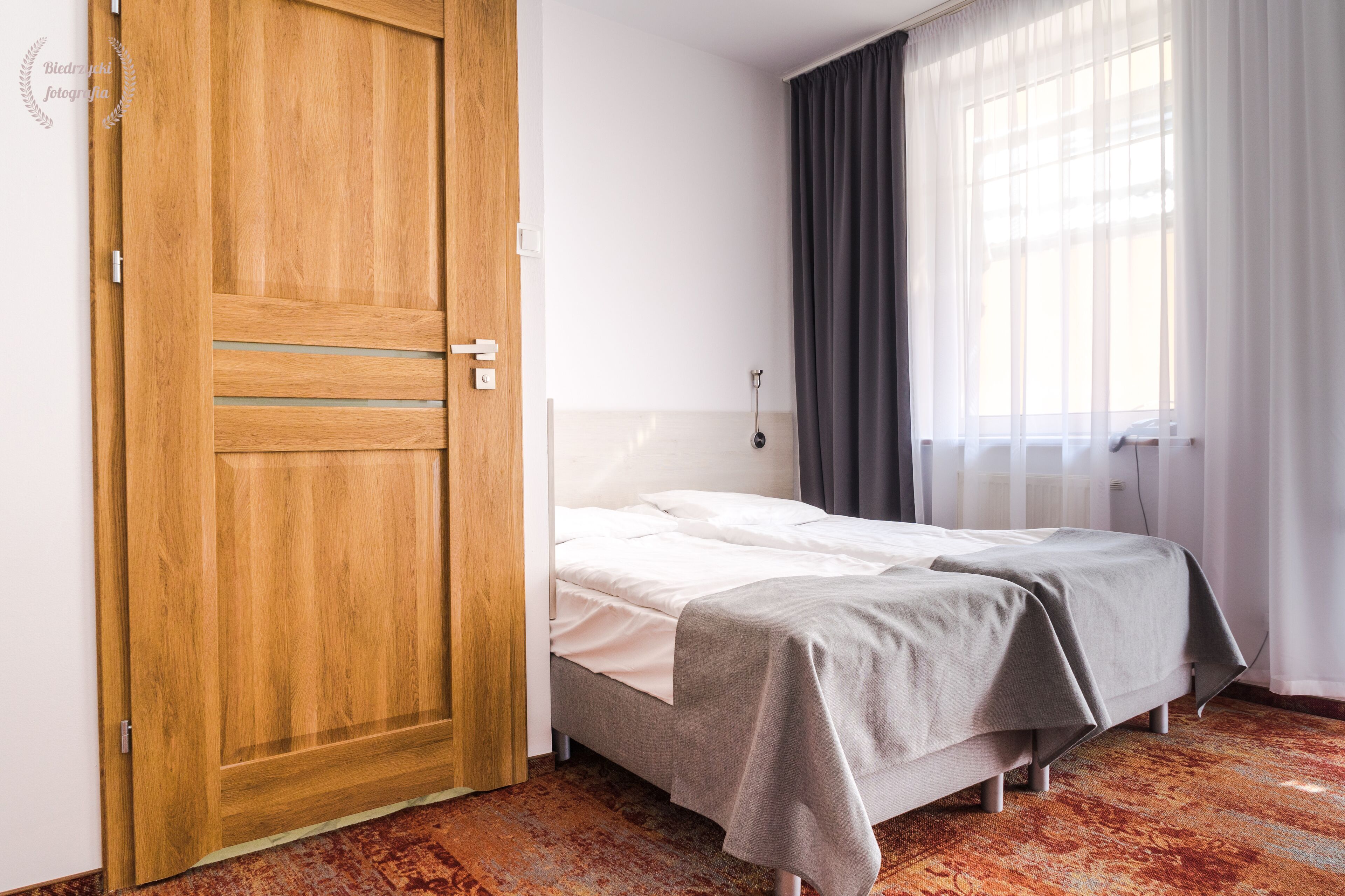 Una habitación de hotel con un armario de madera, una cama con ropa de cama gris y una ventana con cortinas traslúcidas.