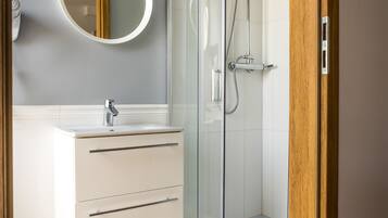 Un baño moderno con ducha envuelta en vidrio, tocador blanco con cajones y un espejo redondo en la pared.
