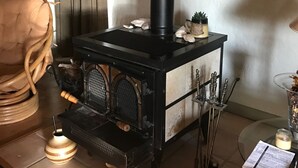 Una estufa de leña con chimenea negra, una cesta tejida y una planta en maceta en una habitación con suelo de madera y una planta grande en la esquina.