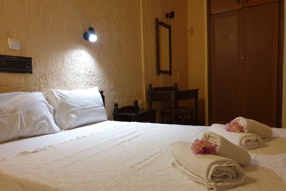 Hotellihuone, jossa on sänky, puinen vaatekaappi, pieni pöytä ja seinälle kiinnitetty peili.