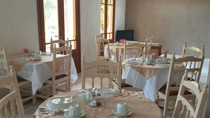 Un comedor con mesas redondas preparadas para cuatro comensales, cada una con mantel blanco, sillas de madera y cubertería ordenada.
