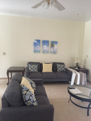 A living room with a sofa, a coffee table, and a ceiling fan. There are three wall-mounted paintings depicting tropical scenes. - Baldwin Camp Rental Properties House #2, is located in North Caicos, TCI (North Caicos)