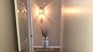 A hallway with wooden flooring, a white door on the left, and a wall-mounted light fixture with a vase of dried branches and flowers. - Wine Country Getaway (Burdett)
