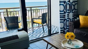 Ein Balkon mit Meerblick, eine Couch, ein Couchtisch mit Blumenvase und ein gemustertes Vorhang.