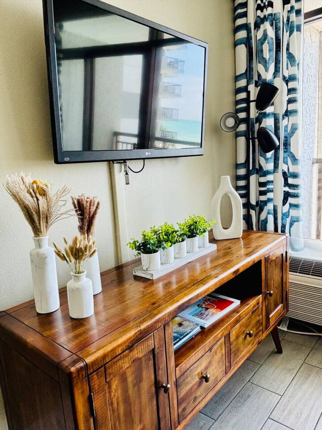 Une console en bois sur laquelle est fixée une télévision à écran plat, un vase contenant des fleurs séchées, une petite plante et un vase blanc.