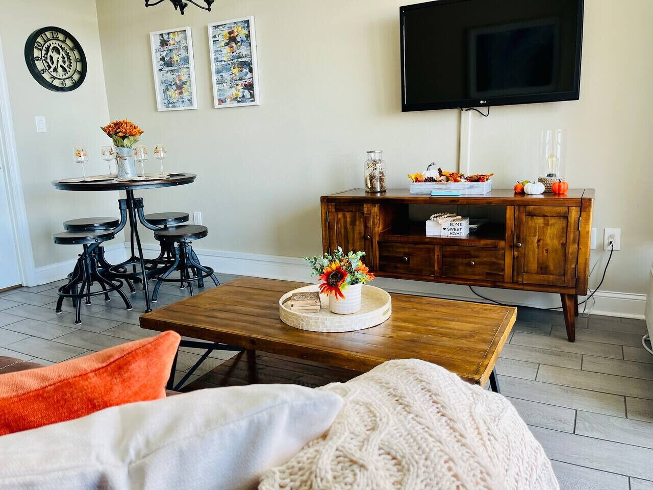 Una sala de estar con una mesa de centro de madera, un televisor montado en la pared, un comedor con una mesa redonda y sillas, y un aparador con objetos decorativos.