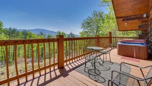 Una terraza de madera con jacuzzi, mobiliario exterior y vistas a montañas.
