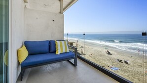 Een balkon met een blauwe bank en een geel kussen, met uitzicht op een strand met mensen en parasols.