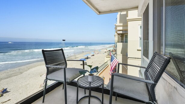 Een balkon met twee stoelen, een kleine tafel en een Amerikaanse vlag. Het balkon heeft uitzicht op een strand, de oceaan en een gebouw op de achtergrond.