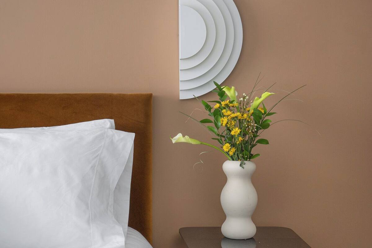 A neatly made bed with white linens, a brown headboard, a white vase with yellow flowers on a black side table, and a wall-mounted circular mirror.