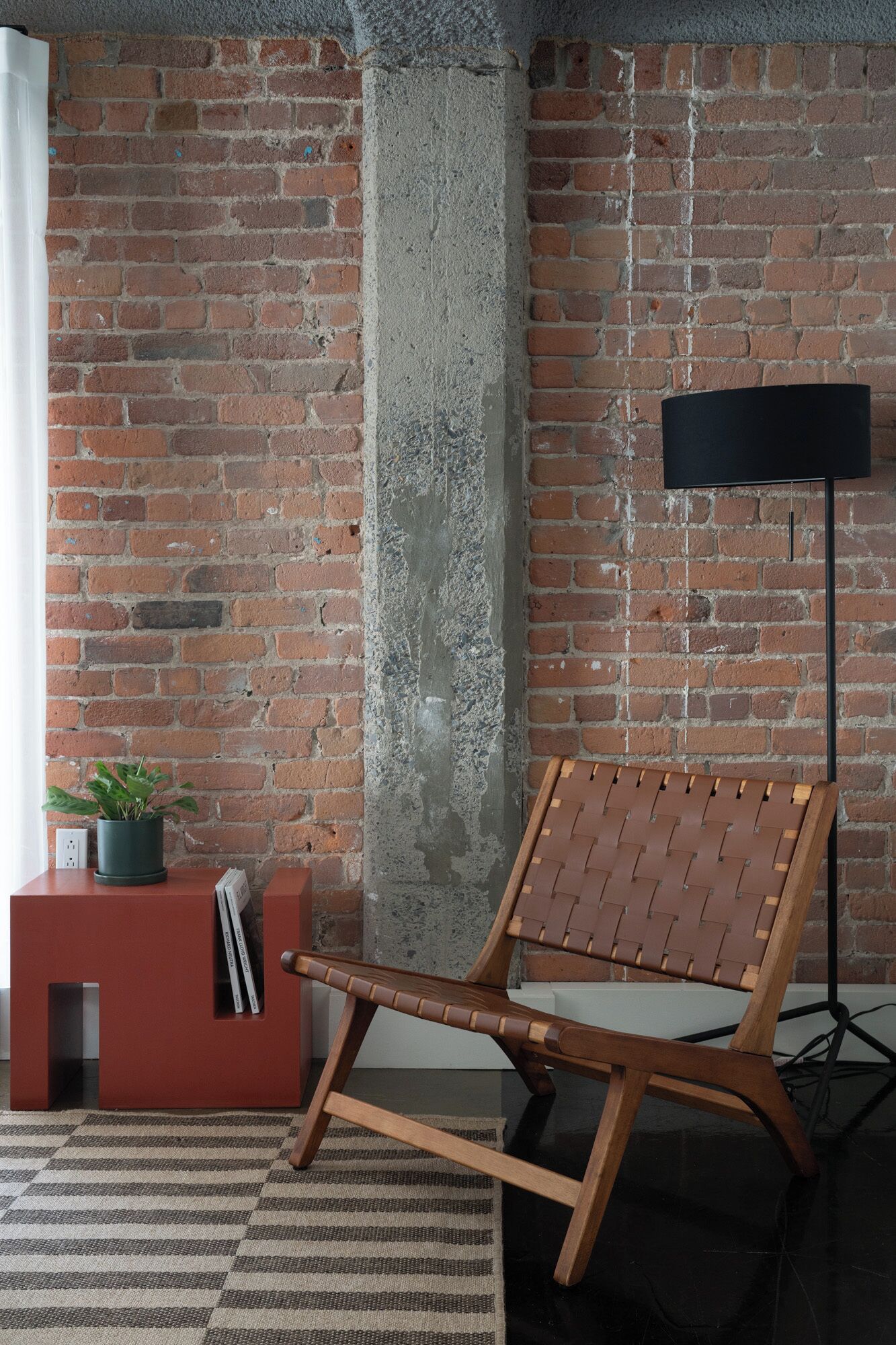 A room with exposed brick walls, a concrete pillar, a wooden chair, a red side table with a plant, and a black floor lamp.