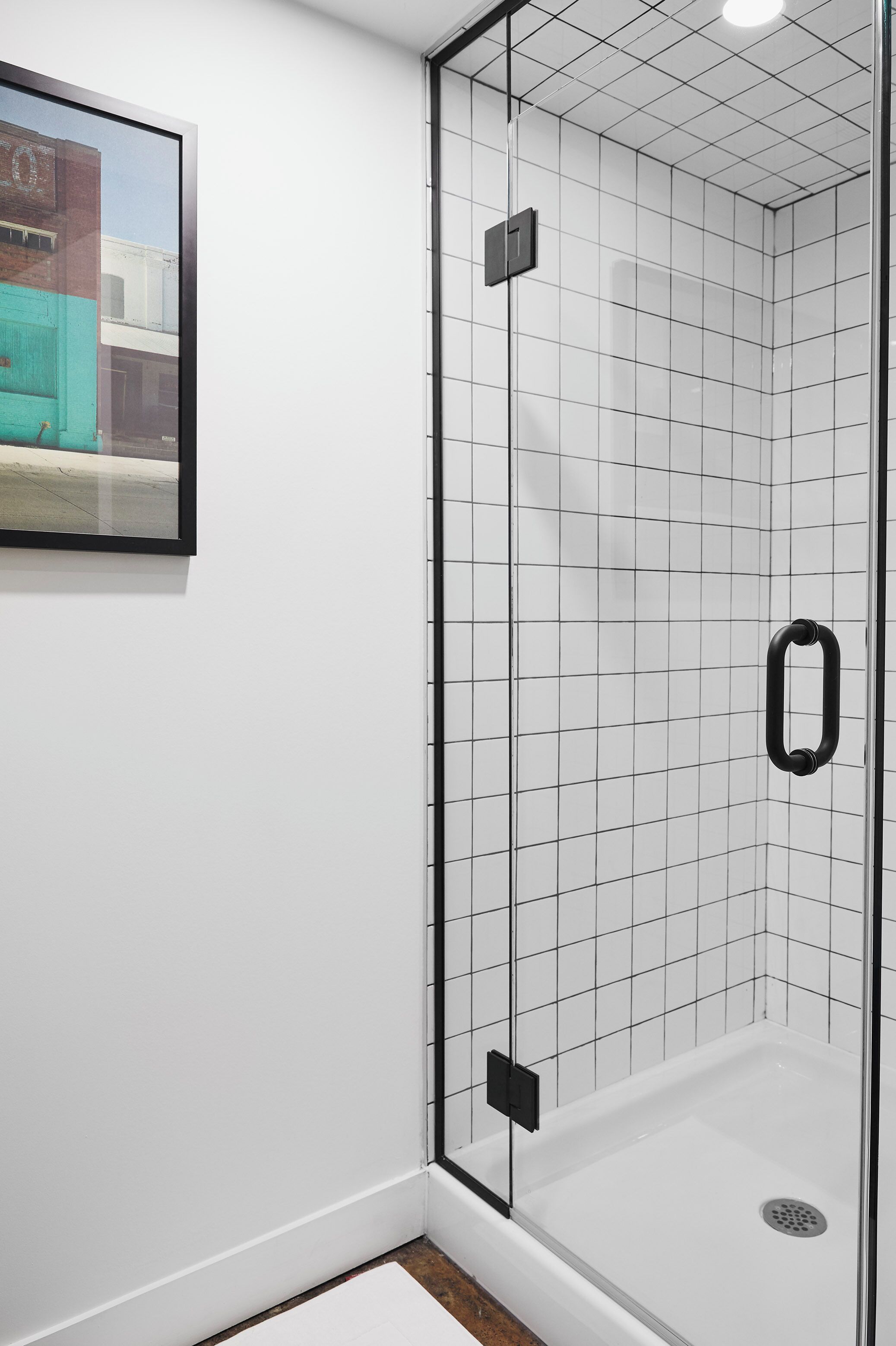 A glass-enclosed shower with a black handle, white tiled walls, and a framed picture on the wall.