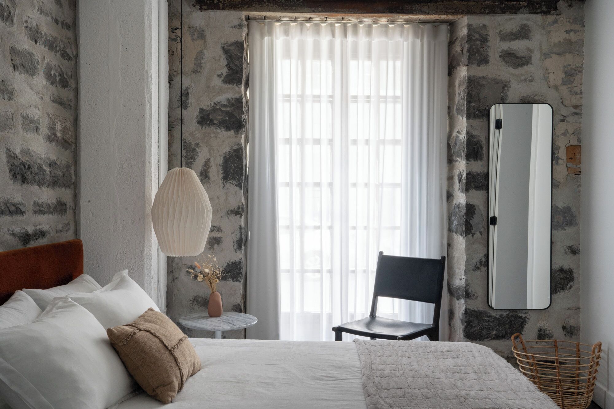 A bedroom with a bed, a chair, a round table with a vase, and a large mirror on the wall.
