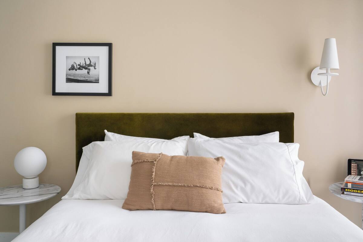 A neatly made bed with white and brown pillows, a framed picture on the wall, a white lamp on a side table, and a small stack of books on a side table.